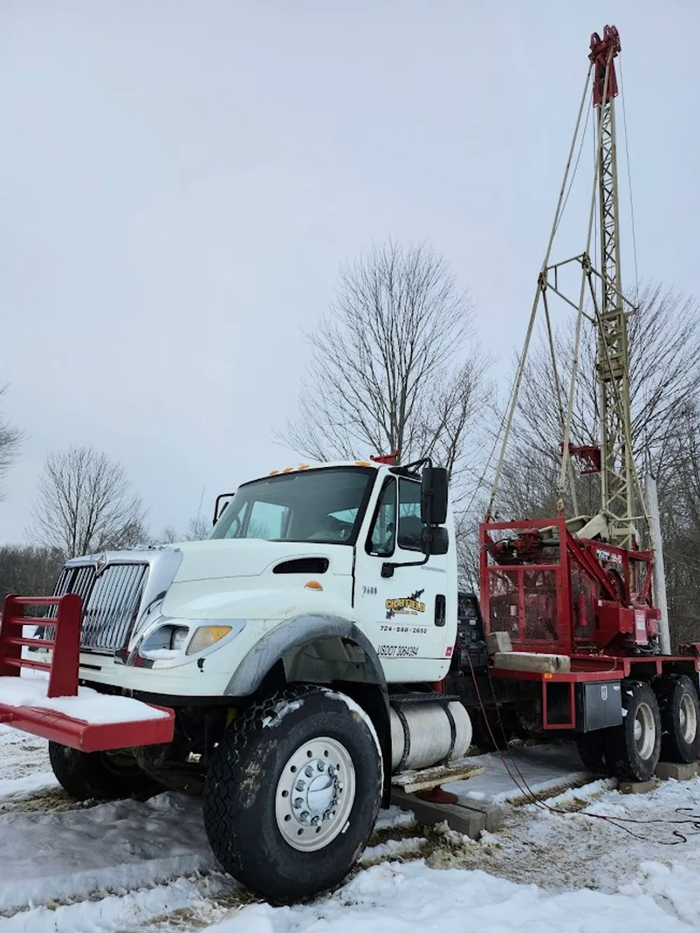 A red Chatfield truck-mounted drilling rig with its mast raised on a snowy road